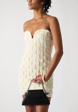 Kremfarget strikket minikjole med v-halsdesign, kombinert med en liten sort skinnhåndveske. Har et sølvfarget kjedehåndtak.