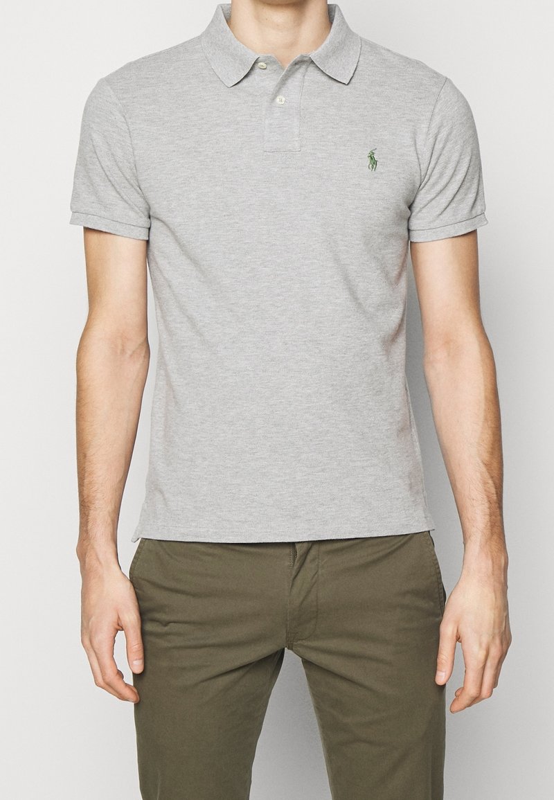 Homme portant un polo gris clair à manches courtes avec une patte de boutonnage à deux boutons et un pantalon vert olive, debout devant un fond uni.