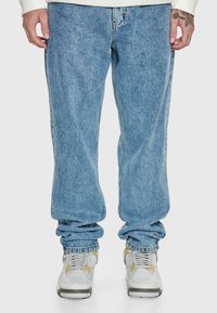 Person wearing loose light blue jeans and white sneakers, standing with hands relaxed by sides on a plain background.
