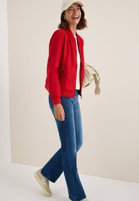 Femme souriante marchant, portant une veste bomber rouge, un jean bleu, des baskets blanches et un chapeau seau beige, tenant un sac beige.