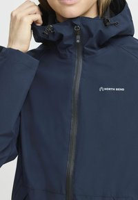 Marineblaue wasserdichte Jacke mit hohem Kragen, verstellbarem Kapuze und Vorderreißverschluss. Hat ein kleines weißes Logo auf der linken Seite.