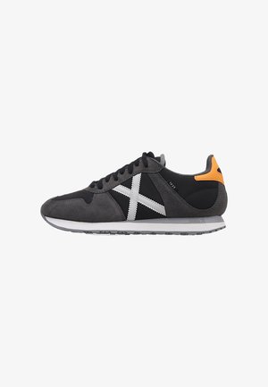 Zapatilla de ante y malla gris con un acento blanco en forma de X, paneles negros, detalle naranja en el talón y suela texturizada. Diseño bajo.