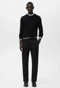 Schwarz strukturierter Pullover, kombiniert mit einem weißen T-Shirt mit Rundhalsausschnitt, dazu eng anliegende schwarze Hosen und schwarze Schuhe. Einfaches und minimalistisches Design.