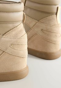 Beige korkeavartiset tennarit, joissa on sileä nahkaverhoilu ja mokkaakkentteja; sisältää ristikkäin ommeltua saumaa ja teksturoidun kumipohjan.
