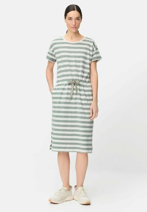 Kvinde stående iført en knælængde, kortærmet, grå- og hvide stribet kjole med snøre i taljen og hvide sneakers.