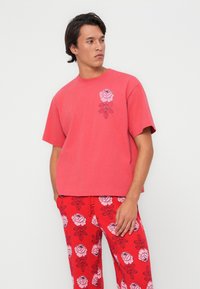 Camiseta de algodón roja de manga corta y cuello redondo, con un gráfico de una rosa rosa en el lado izquierdo del pecho. Combinada con pantalones rojos adornados con un patrón floral.