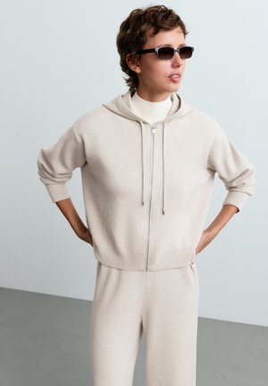 Un mannequin pose, les mains sur les hanches, avec des lunettes de soleil foncées et un sweat à capuche crème assorti, zippé, ainsi qu’un pantalon.