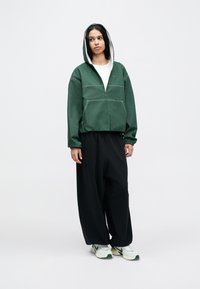 Sweat à capuche vert avec fermeture éclair, doublure blanche et coutures apparentes, associé à un pantalon large noir et des baskets vert clair.
