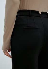 Zwarte broek van een textuurstof, met een achterzak en een subtiele V-vormige tailleband, gecombineerd met een beige lange mouwen top.