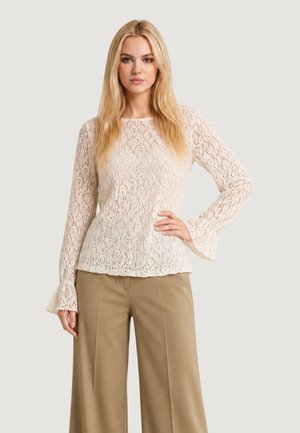 Giovane donna con capelli biondi lunghi che indossa una maglia crema in pizzo a maniche lunghe e pantaloni beige a gamba larga, in piedi con una mano sui fianchi.
