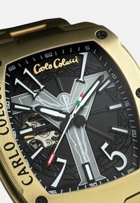 Gouden horloge met een zwarte gestructureerde wijzerplaat, witte en rode accenten, en een uniek kruisontwerp. Beschikt over grote uurmarkeringen en een zichtbaar mechanisme.