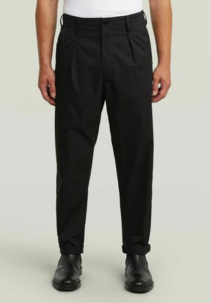 Homme portant un pantalon noir plissé avec des ourlets retroussés et des chaussures en cuir noir, debout devant un fond uni.