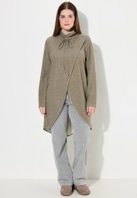 Tunika a maniche lunghe di colore verde oliva con collo a cappuccio e laccio regolabile. Presenta un orlo asimmetrico e un tessuto morbido e leggero. Abbinata a jeans chiari.