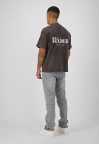 Grijze oversized t-shirt met het "Blekbnns" logo op de achterkant, gecombineerd met lichtblauwe jeans en witte sneakers, weergegeven vanaf de achterkant.