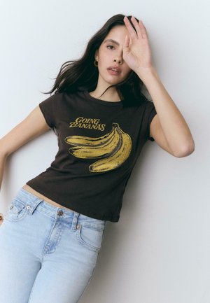 Femme aux cheveux bruns portant un t-shirt noir avec des bananes jaunes et le texte "GOING ZANANAS", associée à un jean bleu clair, levant la main près du visage.