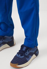 Chaussures de sport bleues avec motifs texturés, semelles intermédiaires blanches, semelles en gomme, portées avec un pantalon bleu vif sur un fond blanc.