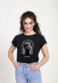 Mladá žena s kudrnatými vlasy, která má na sobě černé tričko s grafikou Wednesday Addams a textem "Nejsem divná, všichni ostatní ano."