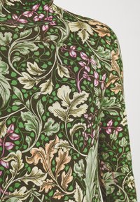 Långärmad topp i mörkgrönt tyg med intrikata blommönster och blad i rosa, creme och grönt, med en mjuk textur.