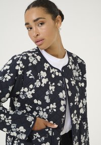 Quilted navy jacket with white floral pattern, round neckline, and front pockets. Features contrast piping along the edges.