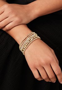 Gouden multistrand armband met getextureerde kralen, gladde metalen accenten en heldere stenen, gedragen om een pols tegen een donkere achtergrond.