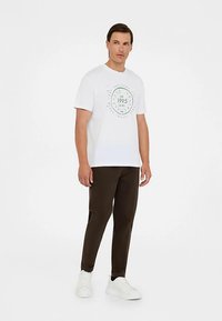 Weißes Baumwoll-T-Shirt mit grünem Kreisgrafik und Text "est. 1995", kombiniert mit dunkelbraunen Hosen und weißen Schuhen, stehend vor einem einfarbigen Hintergrund.