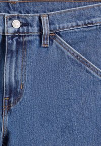 Gros plan sur un jean en denim bleu montrant la poche avant, la passants de ceinture, le bouton en métal et les détails de couture orange.