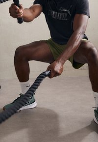 Černé sportovní tričko s logem Puma, zelené šortky, šedé a zelené tenisky, drží provaz. Viditelné silné svaly na nohách a rukou. Neutrální pozadí.