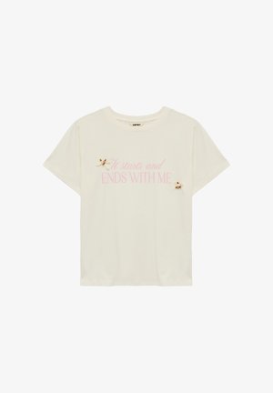 T-shirt en coton crème à manches courtes. Présente un texte rose "Ça commence et se termine AVEC MOI" et deux petites décorations florales sur le devant.