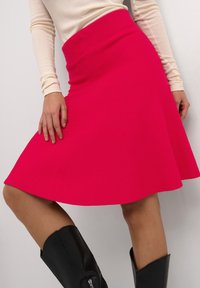 Jupe évasée rose vif avec une large ceinture, fabriquée en tissu texturé, assortie à des bottes noires jusqu'aux genoux.