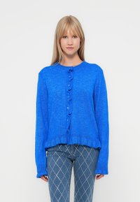 Cardigan en tricot bleu avec un col rond, présentant une fermeture à boutons et un bord ondulé. Les manches sont longues avec des poignets côtelés.