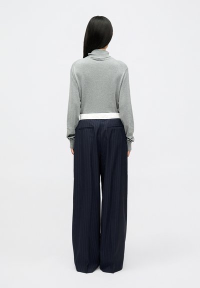 Pelēks džemperis ar augstu apkakli, apvienots ar zilām biksēm ar svītrām un baltu jostasvietu.