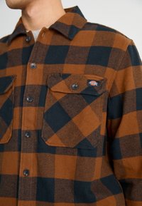 Camisa de franela con cuadros marrones y negros, con cierre de botones, dos bolsillos frontales y un pequeño parche con logo en el bolsillo izquierdo.