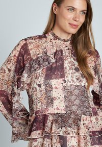 Bluse mit Blumenmuster in einer Mischung aus Burgunder- und Pfirsichtönen. Verfügt über Rüschenschößen und einen hohen Ausschnitt, hergestellt aus leichtem Stoff.