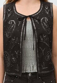 Zwarte mouwloze vest met zilveren pailletten en kraaltjes in paisleypatronen. Vetersluiting aan de voorkant en een lichtgrijze top eronder met een textuur.