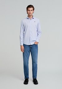 Gestreiftes, hellblaues Hemd mit langen Ärmeln, kombiniert mit mittelblauen Jeans und schwarzen Schuhen. Verfügt über ein kleines Logo auf der Brust.
