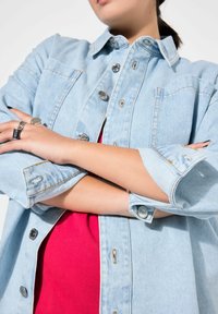Veste en denim bleu clair avec fermeture à boutons, poches poitrine et manches retroussées, superposée sur une chemise rouge. Comprend des coutures et des accessoires visibles.