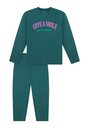 Smilende pige iført teal sweatshirt og bukser med teksten "GIVE A SMILE get a smile,", der viser biceps med løftede arme, barfodet på hvid baggrund.