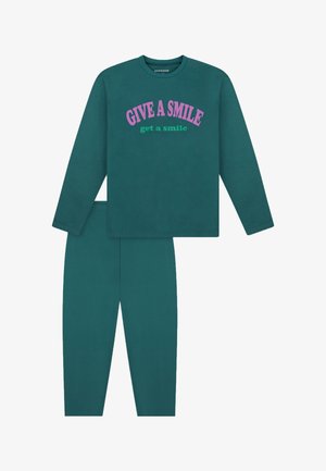 Smilende pige iført teal sweatshirt og bukser med teksten "GIVE A SMILE get a smile,", der viser biceps med løftede arme, barfodet på hvid baggrund.
