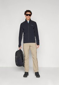 Black zip-up jacket, beige pants, black backpack, and black shoes. Model wearing dark sunglasses, stands against a plain background.