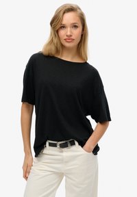 Camiseta negra holgada con un escote ancho y mangas cortas, combinada con pantalones de color claro y un cinturón decorativo.