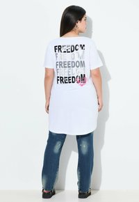 Camiseta blanca de algodón con la palabra "FREEDOM" impresa repetidamente en negro y plata en la parte de atrás. Combinada con jeans desgastados azules y zapatos negros.