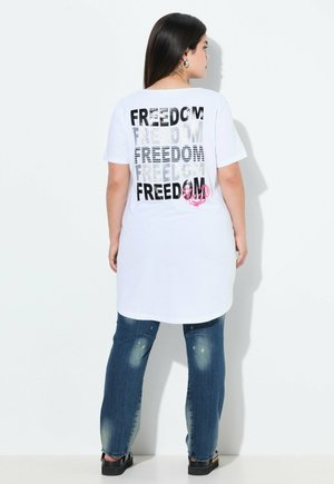 Witte katoenen T-shirt met "FREEDOM" herhaaldelijk in zwart en zilver gedrukt op de rug. Gecombineerd met blauwe versleten jeans en zwarte schoenen.