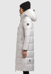 Svetle šedý péřový kabát s kapucí, prošívaný design, boční kapsy a černá nášivka loga na rukávu. Kombinováno s černou pletenou čepicí.