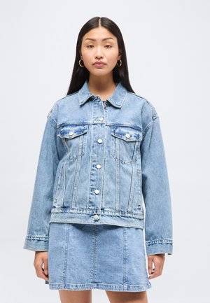 Nő világoskék farmerkabátot visel gombolással és hozzáillő farmer szoknyával, egyszerű világos háttér előtt áll.