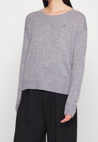 Pull en tricot gris avec un ourlet et des poignets côtelés. Présente un petit logo sur la poitrine. Coupe décontractée, encolure bateau et tissu texturé.