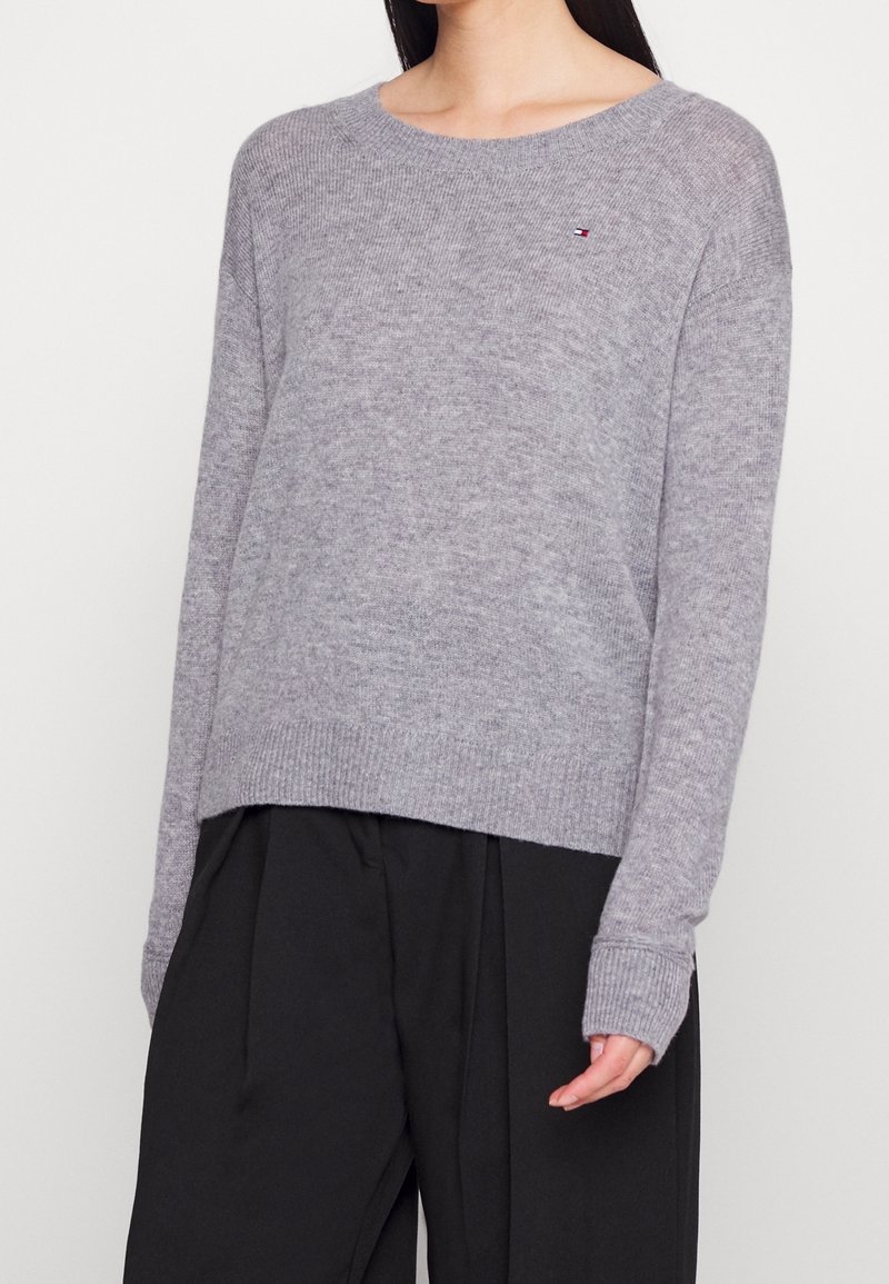 Pull en tricot gris avec un ourlet et des poignets côtelés. Présente un petit logo sur la poitrine. Coupe décontractée, encolure bateau et tissu texturé.