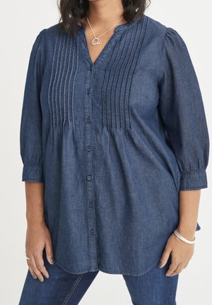 Kvinde iført en mørkeblå knap-op bluse med lange ærmer med lodrette plisser og mørkeblå jeans, accessoriseret med sølvhalskæde, ringe og armbånd.
