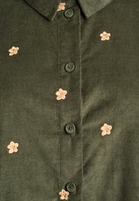 Grünes Cordhemd mit beiger Blumenstickerei. Auffällige Details sind der spitze Kragen und die durchgeknöpfte Vorderseite mit grünen Knöpfen.