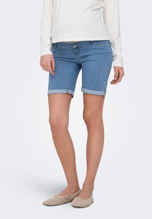 Femme portant un short en jean bleu clair retroussé, un haut blanc à manches longues et des chaussures beige tissées à enfiler, debout devant un fond uni.