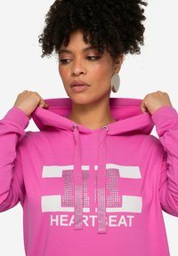 Sudadera rosa con un gráfico blanco que presenta la palabra "HEARTBEAT" y adornos. Cuenta con una capucha con cordón y una textura suave de mezcla de algodón.